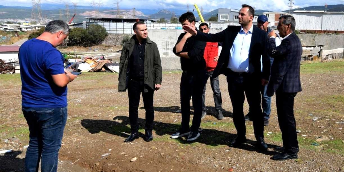 Açık havada, toprak ve seyrek çimlerle kaplı geniş bir alanda duran altı kişilik bir grup erkek, bir projeyi veya araziyi inceliyor. Gruptan biri elini ileriye doğru uzatarak bir noktayı işaret ediyor ve yanındakilerle konuşuyor. Diğerleri ise dikkatle onu dinliyor veya çevreyi gözlemliyor. Grubun solunda mavi tişörtlü bir kişi elindeki tablete veya telefona bakarken, diğerleri ceket ve gömlek gibi günlük kıyafetler giymiş durumda. Arka planda ise sanayi yapıları, elektrik direkleri ve uzaklarda dağlık bir arazi görülüyor.