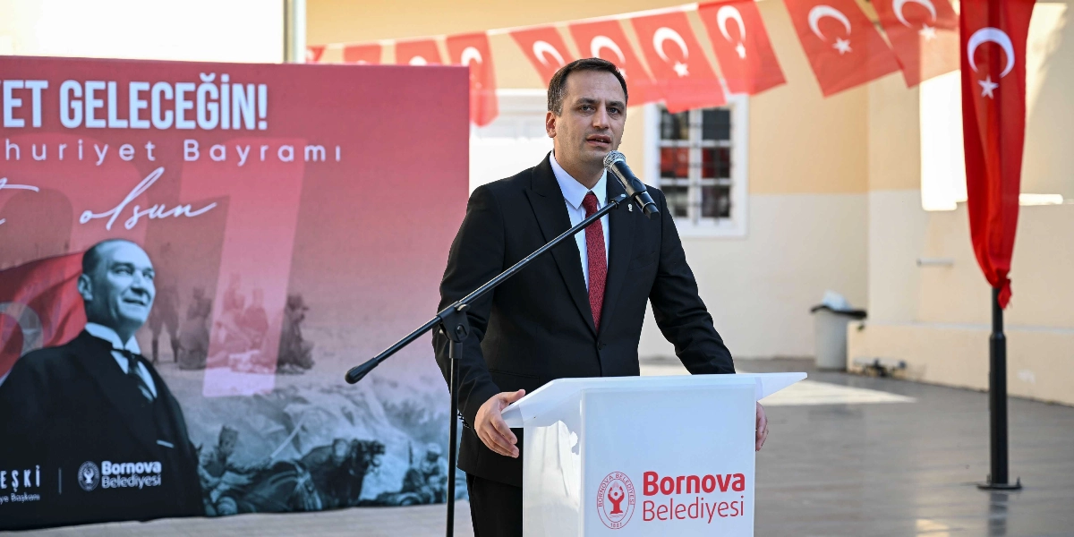 Koyu renk takım elbiseli ve kırmızı kravatlı bir erkek, üzerinde Bornova Belediyesi logosu bulunan beyaz bir kürsüde mikrofon başında konuşma yapıyor. Arka planda Mustafa Kemal Atatürk'ün görselinin ve 