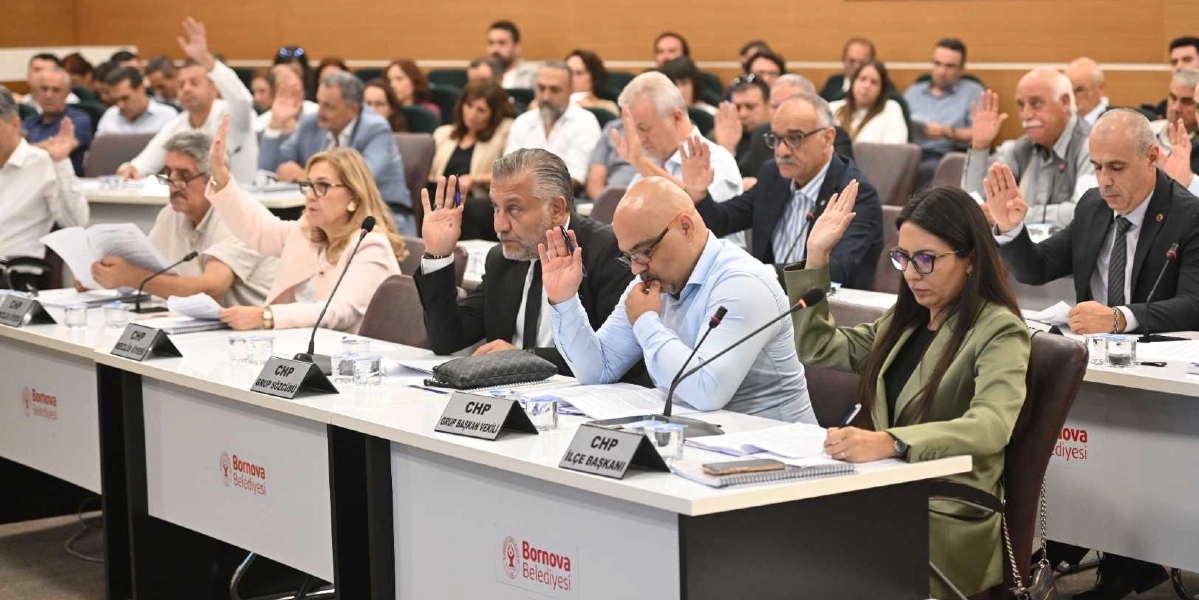 Bornova Belediyesi meclis salonunda, masalarda oturan çok sayıda meclis üyesi ellerini havaya kaldırarak oy kullanıyor. Ön planda, üzerinde 
