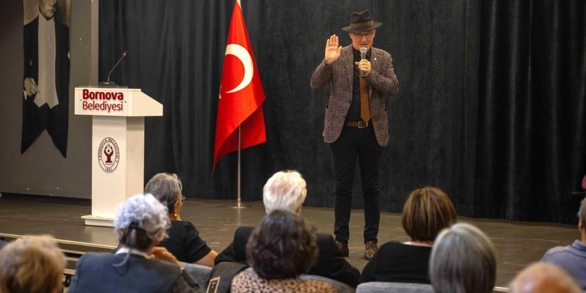 Siyah perdeli bir sahnede, üzerinde Bornova Belediyesi logosu ve yazısı bulunan kürsünün yanında, fötr şapkalı, kahverengi ceketli ve kravatlı bir konuşmacı elinde mikrofonla dinleyicilere hitap ediyor. Konuşmacı sağ elini havaya kaldırmış durumda ve arkasında Türk bayrağı ile Atatürk'ün siyah beyaz bir portresi yer alıyor. Ön planda ise söyleşiyi takip eden izleyicilerin sırtları görünüyor.