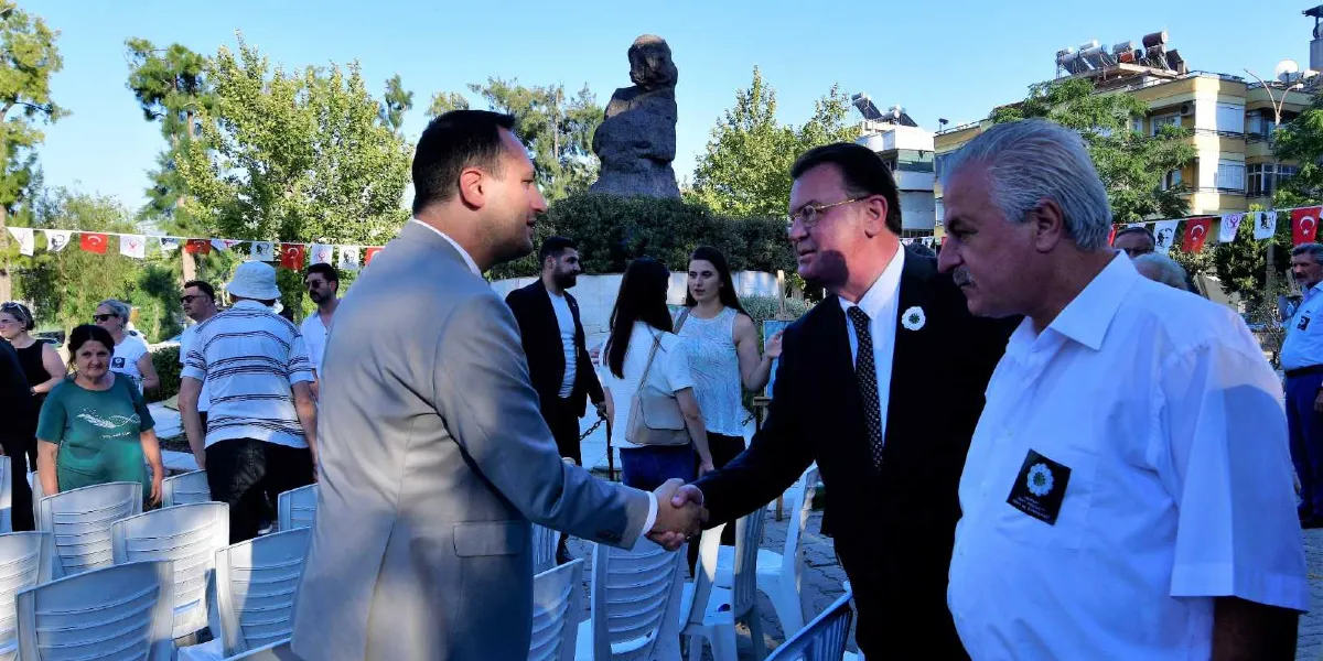Açık havada düzenlenen bir anma töreninde, gri takım elbiseli bir erkek ile koyu renk takım elbiseli ve kravatlı bir erkek tokalaşıyor. Sağ tarafta, üzerinde Srebrenica anma çiçeği figürlü bir yaka kartı bulunan beyaz gömlekli bir adam onları izliyor. Arka planda boş beyaz plastik sandalyeler, ağaçlar, bir anıt heykeli ve Türk bayraklarıyla süslenmiş bir alan görülüyor.