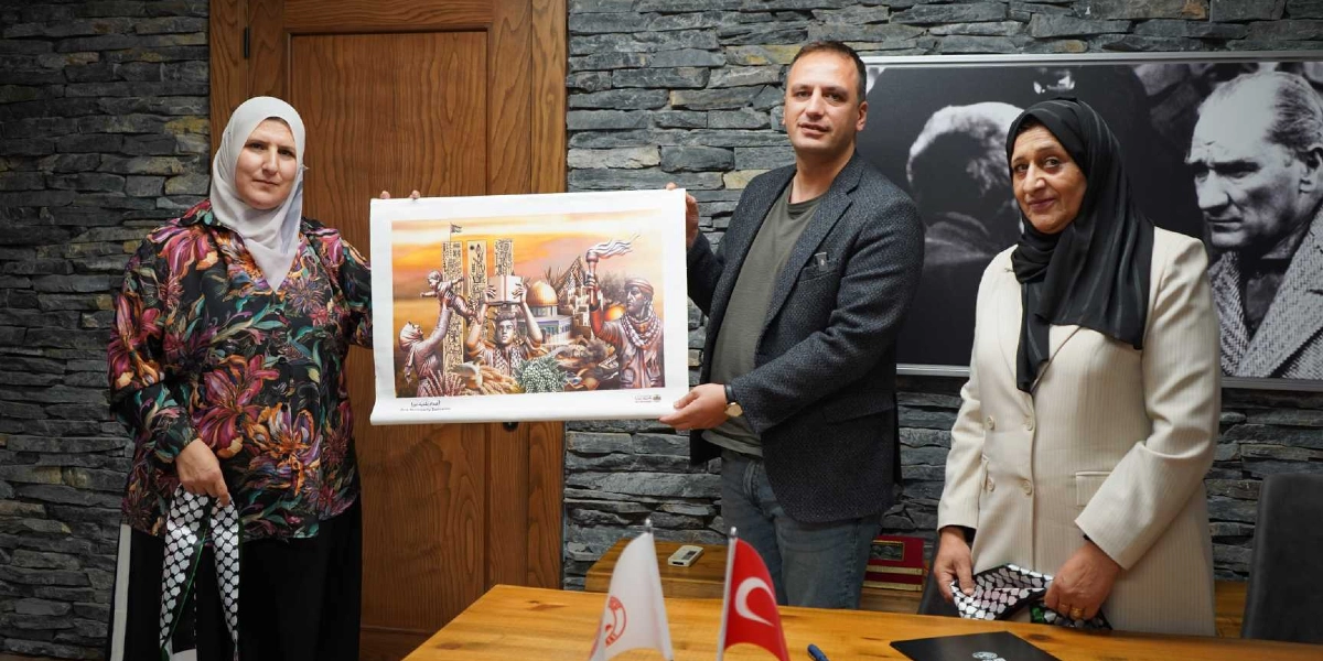 İki kadın ve bir erkek, ellerinde Filistin'i simgeleyen bir tabloyu birlikte tutarak poz veriyor. Soldaki kadın beyaz başörtüsü ve desenli bir bluz giyiyor, sağdaki kadın ise siyah başörtüsü ve krem rengi bir ceket giyiyor. Ortadaki erkek ise gri bir tişört ve üzerine koyu renkli bir ceket giymiş durumda. Arka planda taş duvar üzerinde Mustafa Kemal Atatürk'ün bir portresi yer alıyor. Masanın üzerinde ise küçük bir Türk bayrağı ve bir belediye flaması bulunuyor.
