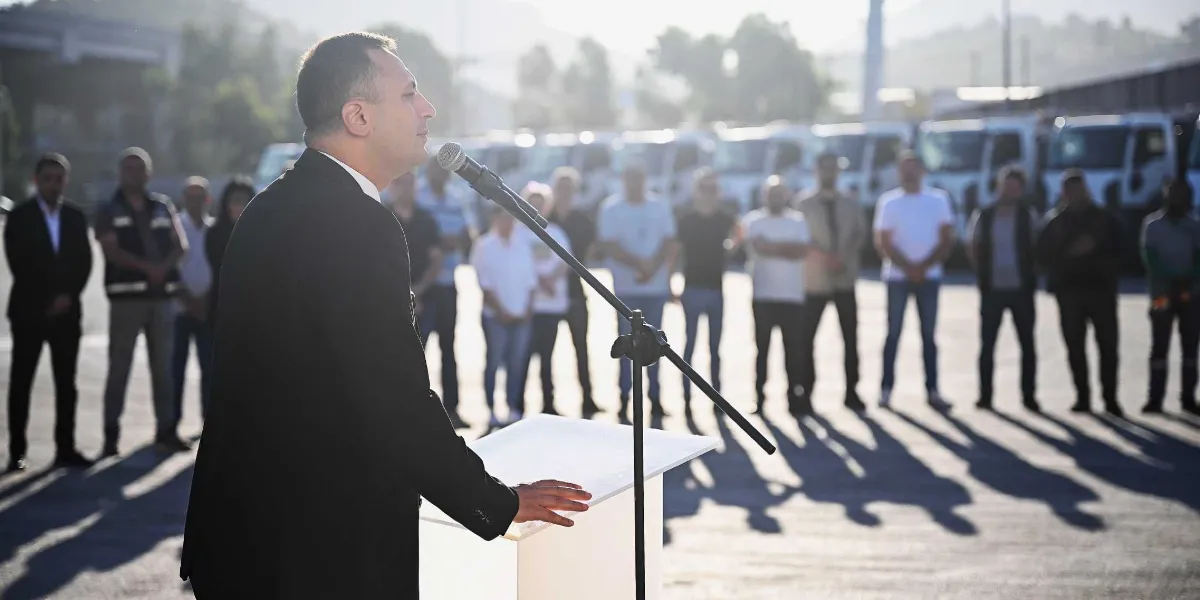 Koyu renk ceket giymiş bir erkek, açık havada bir kürsüde mikrofon başında konuşma yapıyor. Arka planda, bir otopark alanında sıralanmış beyaz kamyonların önünde duran ve konuşmacıyı dinleyen bir grup insan bulunuyor.