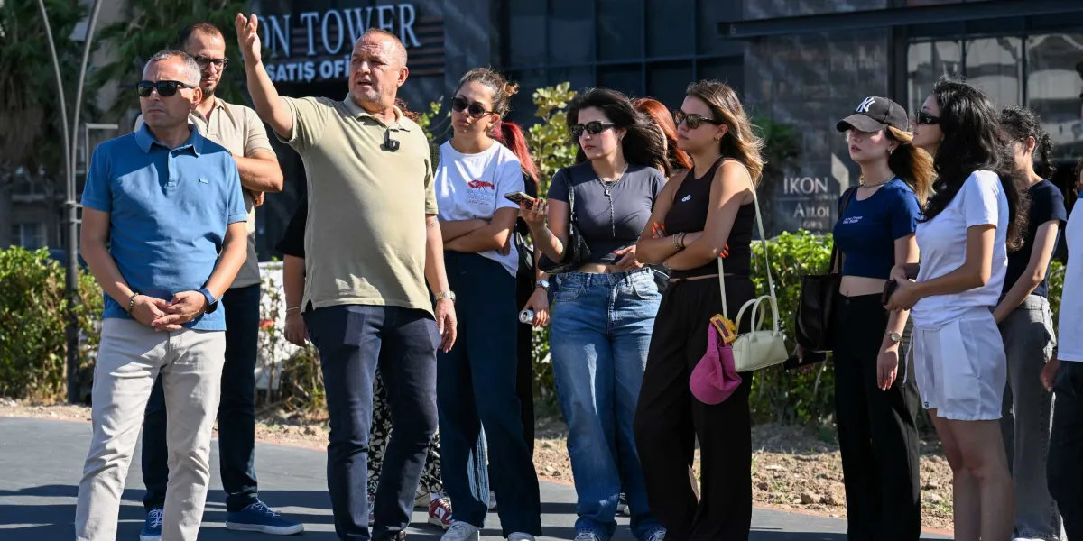 Açık havada, Ikon Tower satış ofisinin önünde duran ve bir şeyler dinleyen bir grup insan. Grubun önünde duran orta yaşlı bir erkek, eliyle bir yönü işaret ederek konuşuyor. Gruptaki diğer kişiler, çoğunluğu genç kadınlardan oluşan, günlük kıyafetler ve güneş gözlükleri takmış bir şekilde dikkatle onu dinliyor.