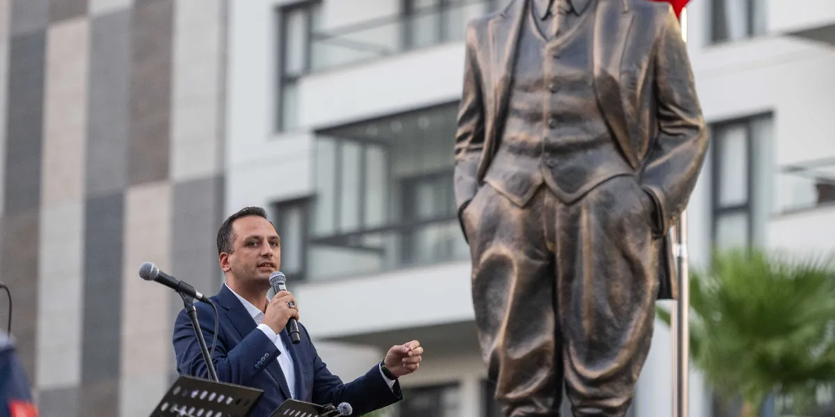 Koyu renk takım elbiseli bir erkek, elinde mikrofonla konuşma yaparken görülüyor. Arka planda ise modern bir apartman binası ve bronz renkli, elleri pantolonunun cebinde duran bir Atatürk heykeli yer alıyor.