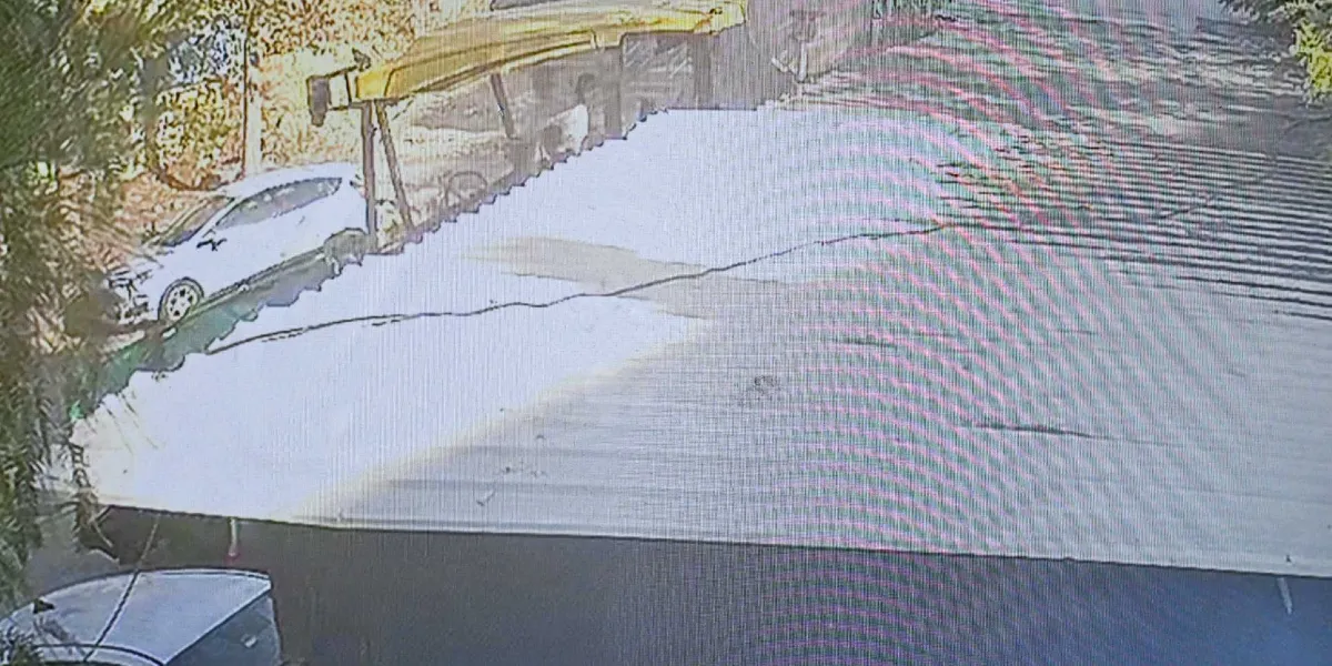 Güvenlik kamerası açısından çekilmiş, düşük çözünürlüklü görüntüde, bir otopark alanında park halindeki beyaz bir otomobil ve hemen yanında, ön kısmı hasar görmüş veya parçalanmış başka bir araç görülüyor. Araçların üzerinde, gölgelik görevi gören metal bir çatı yapısı bulunuyor.