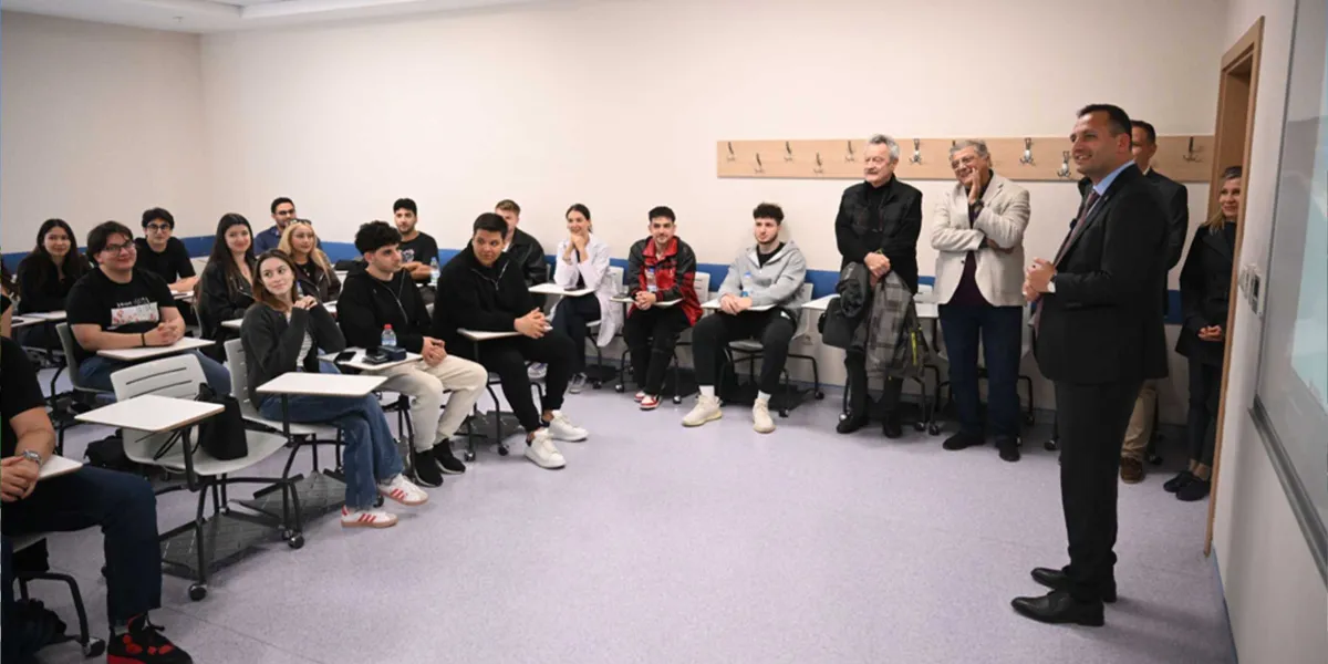 Modern bir sınıfta, takım elbiseli bir konuşmacıyı dikkatle dinleyen gençlerden oluşan bir grup öğrenci ve yanlarında duran yetişkinler. Öğrenciler sıralarında otururken, konuşmacı sınıfın önünde ayakta durarak onlara hitap ediyor.