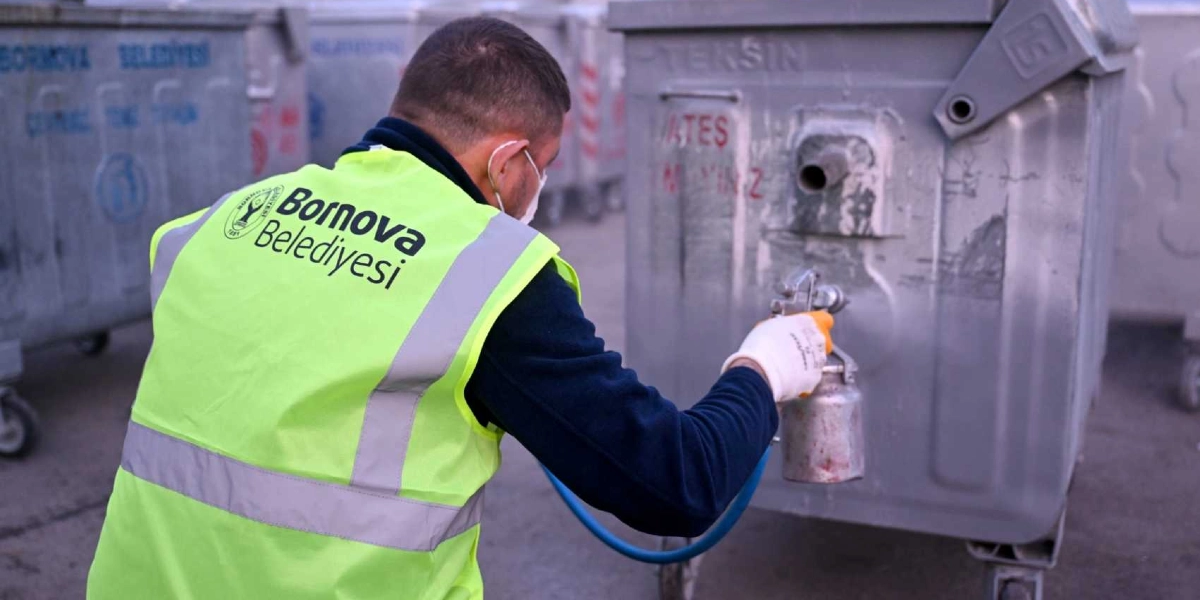 Sırtında Bornova Belediyesi yazılı fosforlu sarı iş yeleği giyen bir belediye çalışanı, elindeki boya tabancasıyla gri renkli metal bir çöp konteynerini boyuyor. Arka planda benzer şekilde sıralanmış başka çöp konteynerleri bulunuyor.
