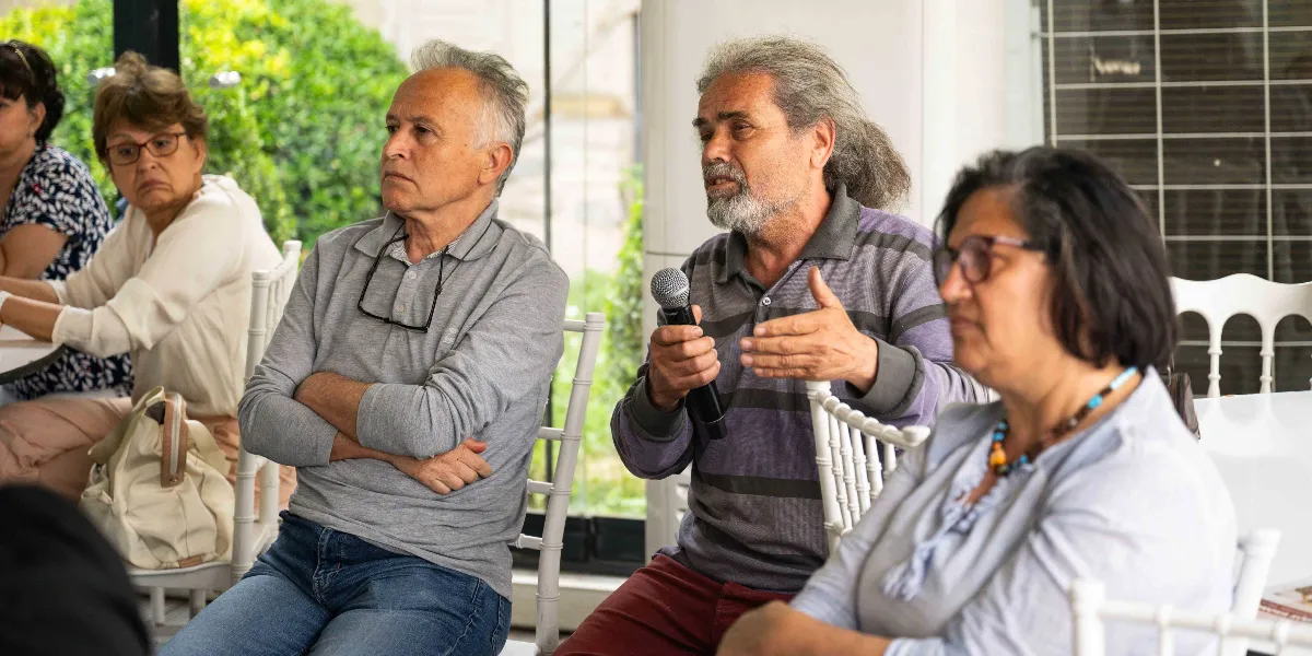 Kapalı bir mekanda düzenlenen bir toplantıda, elinde mikrofon tutan gri çizgili kazaklı ve uzun saçlı bir adam konuşma yapıyor. Yanında gri polo yaka tişörtlü, kolları kavuşturulmuş bir adam ve gözlüklü bir kadın onu dikkatle dinliyor. Arka planda ise başka katılımcılar yer alıyor.