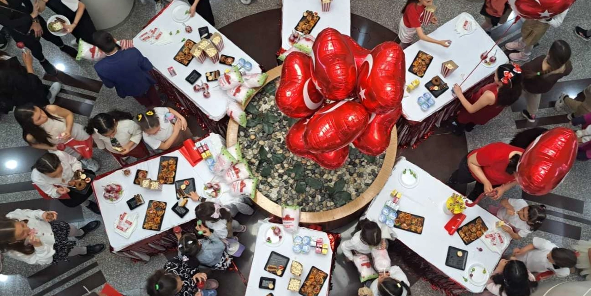 Üstten çekilmiş bir açıyla, bir merkezdeki dairesel taşlı havuzun etrafına yerleştirilmiş beyaz örtülü masalarda 23 Nisan kutlaması yapan çocuklar görülüyor. Masaların üzerinde patlamış mısır kutuları, çeşitli atıştırmalıklar ve içecekler bulunuyor. Havuzun ortasında, üzerinde Türk bayrağı baskısı olan çok sayıda kırmızı kalp şeklinde balon bir arada duruyor. Çocuklar masaların etrafında toplanmış, yiyeceklerini alıyor ve birbirleriyle vakit geçiriyor.