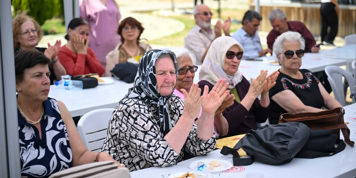 Açık havada düzenlenen bir etkinlikte, masalarda oturan ve alkışlayarak bir kutlamaya katılan ağırlıklı olarak yaşlı kadınlardan oluşan bir grup. Ön planda, başında siyah beyaz desenli bir eşarp bulunan yaşlı bir kadın ellerini çırparak gülümserken, yanında güneş gözlüklü ve başörtülü diğer katılımcılar da ona eşlik ediyor. Masaların üzerinde tabaklar ve su şişeleri bulunuyor, arka planda ise etkinliği izleyen diğer katılımcılar yer alıyor.