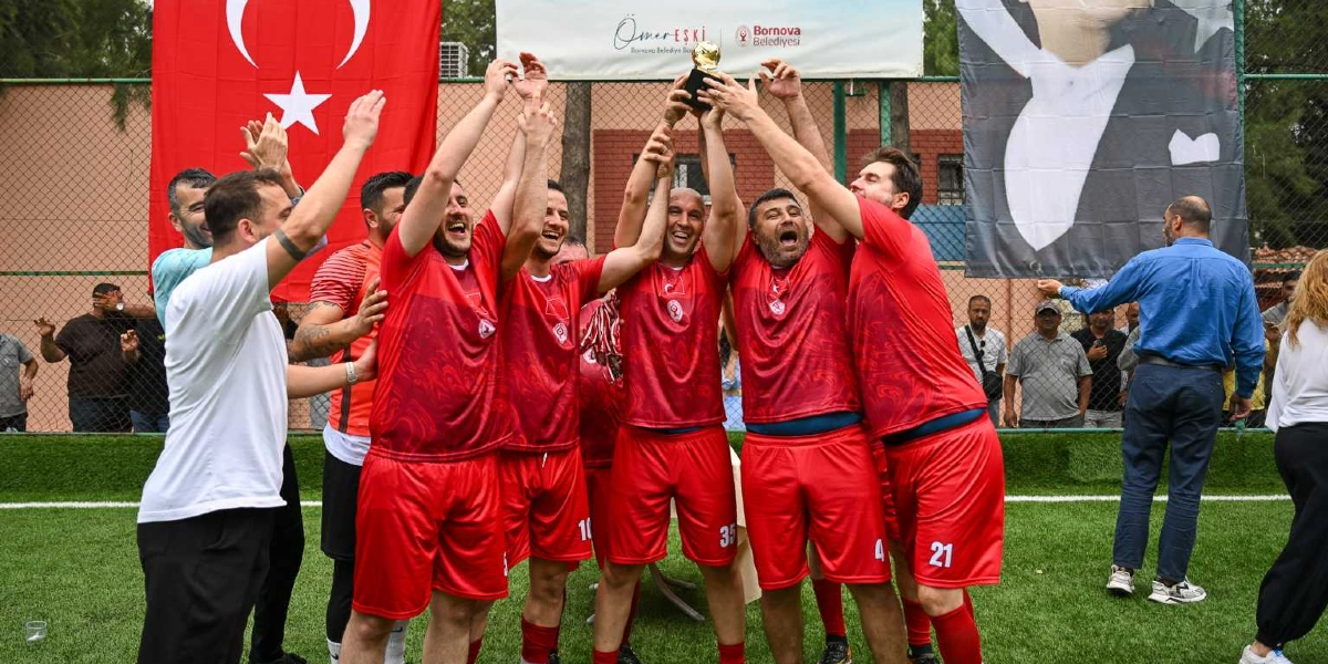 Kırmızı formalar giymiş bir grup futbolcu, halı sahada kazandıkları altın rengi kupayı hep birlikte havaya kaldırarak zaferlerini kutluyor. Oyuncuların arkasında büyük bir Türk bayrağı, Bornova Belediyesi'ne ait bir pankart ve Mustafa Kemal Atatürk'ün silüetinin yer aldığı bir afiş bulunuyor. Çevredeki diğer kişiler de bu coşkulu anı izliyor.