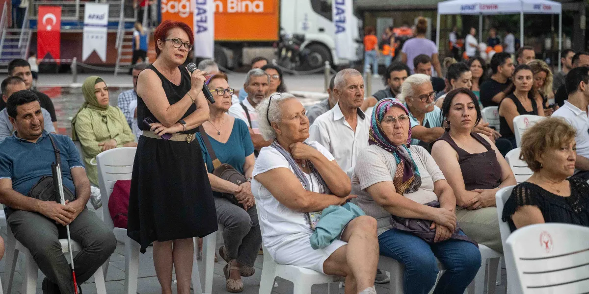 Açık havada düzenlenen bir toplantıda, kısa kızıl saçlı ve gözlüklü bir kadın elinde mikrofonla konuşma yaparken, önünde beyaz plastik sandalyelerde oturan farklı yaş gruplarından katılımcılar onu dinliyor. Arka planda üzerinde AFAD logoları bulunan bir araç ve beyaz çadırlar yer alıyor.