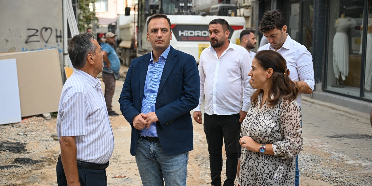 Bornova Belediye Başkanı Mustafa İduğ ve beraberindeki heyet, devam eden yol yenileme çalışmalarını yerinde incelemek üzere bir sokakta incelemelerde bulunuyor. Arka planda üzerinde T.C. Bornova Belediyesi yazılı bir iş makinesi ve çalışma alanı görülüyor.
