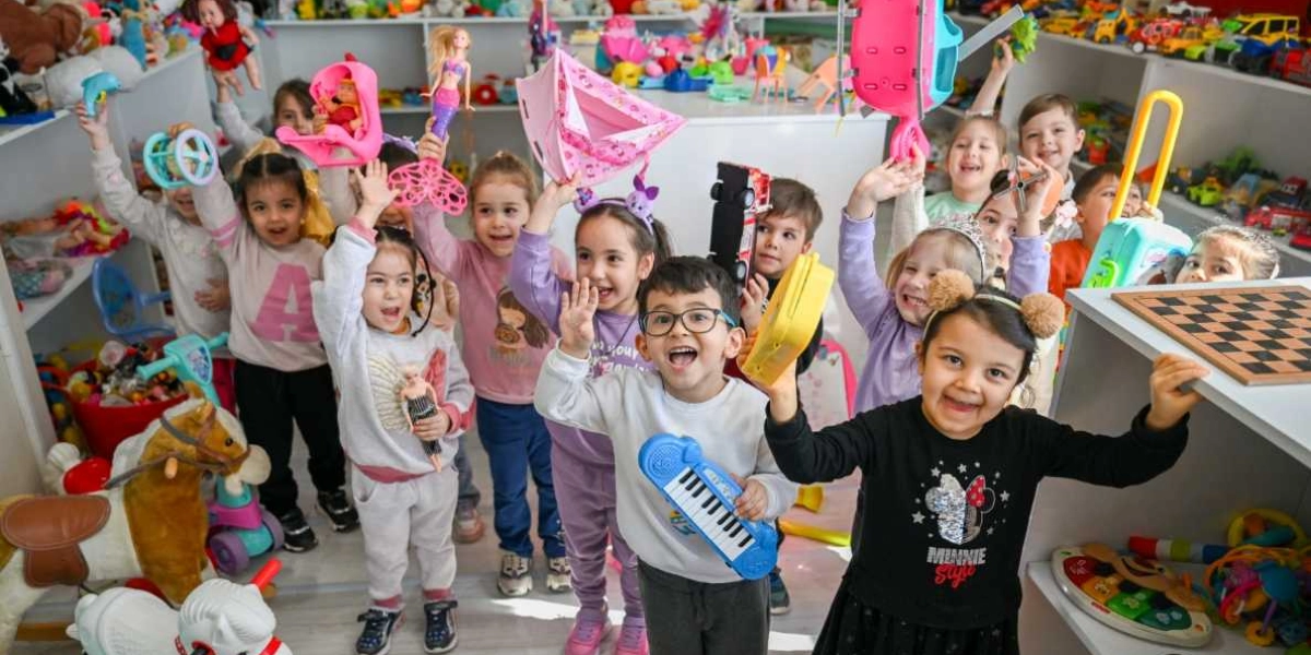Oyuncaklarla dolu bir odada ellerinde çeşitli oyuncaklar tutarak neşeyle poz veren bir grup çocuk. Çocukların bir kısmı ellerinde bebekler, oyuncak piyano, pembe bir çadır ve farklı oyuncaklar tutarken, ön planda gözlüklü bir erkek çocuk ve Minnie Mouse baskılı siyah bir tişört giyen bir kız çocuğu gülümseyerek kameraya bakıyor. Arka planda raflara dizilmiş çok sayıda oyuncak yer alıyor.