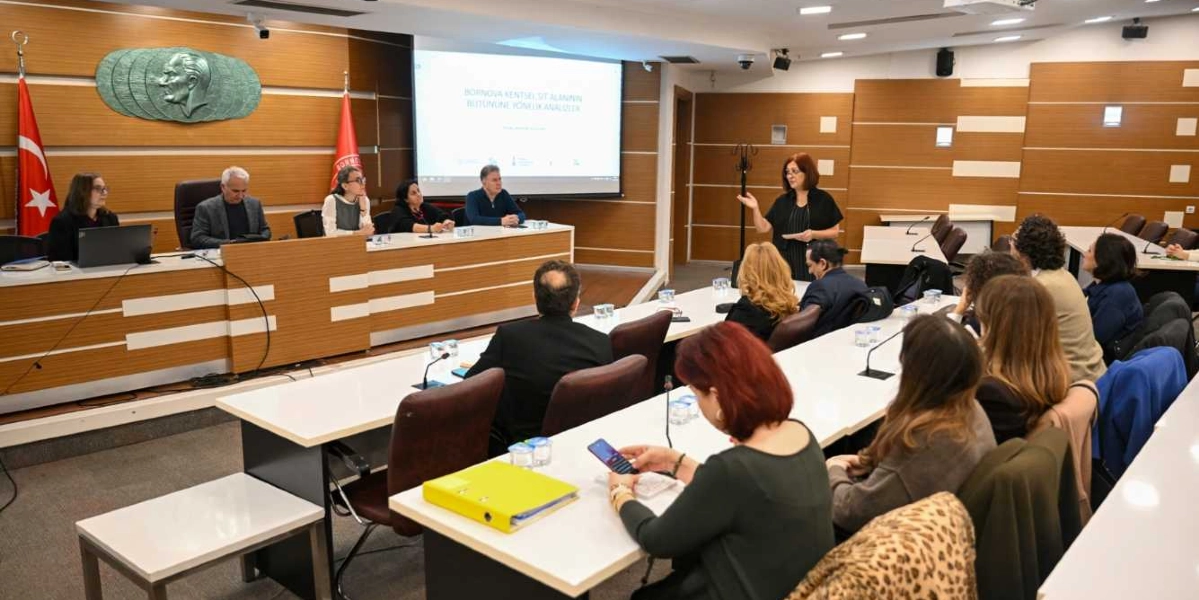 Modern bir toplantı salonunda, Bornova'nın tarihi kimliğine yönelik yol haritasının tartışıldığı bir sunum yapılıyor. Salonun ön tarafındaki masada beş kişilik bir heyet otururken, ayakta duran bir kadın katılımcılara hitap ediyor. Arka planda duvarda Atatürk kabartması, Türk bayrağı ve Bornova Belediyesi'nin flaması yer alıyor. Salonun geri kalanında ise masalarda oturan ve sunumu dinleyen çok sayıda katılımcı bulunuyor.