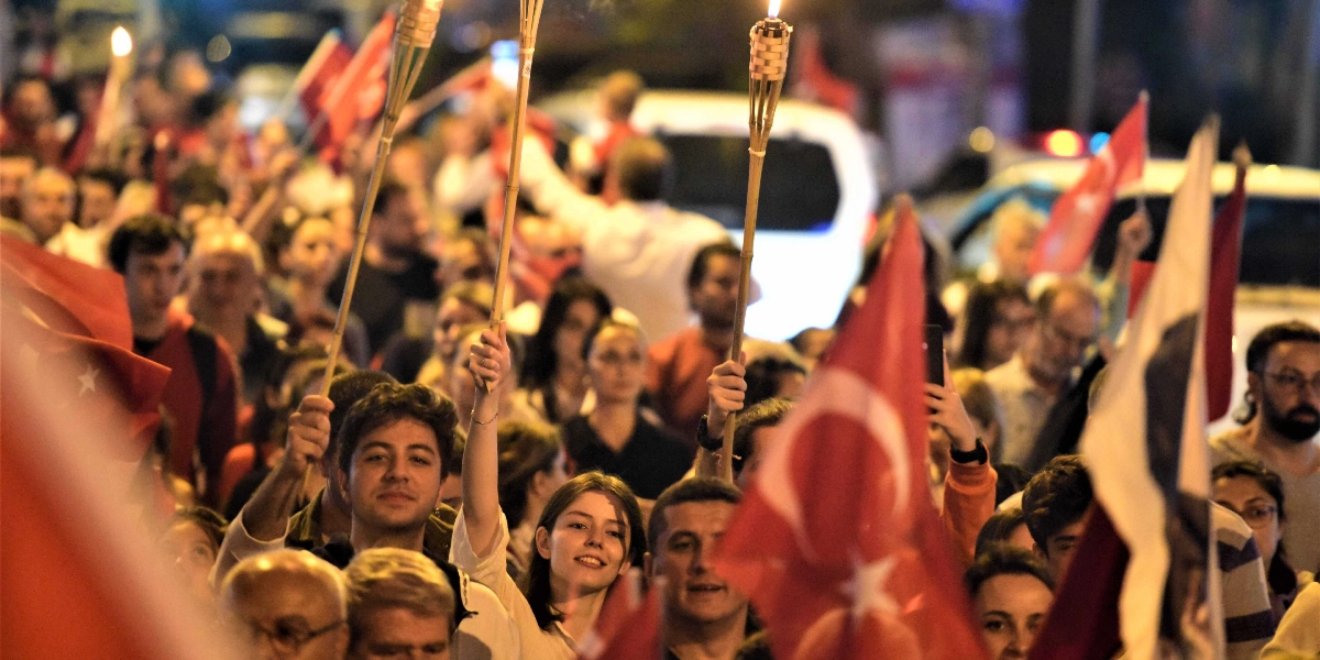 Gece vakti düzenlenen bir fener alayı yürüyüşünde, ellerinde yanan meşaleler ve Türk bayrakları taşıyan kalabalık bir grup insan. İnsanlar neşeli bir ifadeyle yürürken, ön planda meşale tutan genç bir kadın ve erkek dikkat çekiyor. Arka planda ise yürüyüşe katılan çok sayıda kişi ve araç ışıkları seçiliyor.