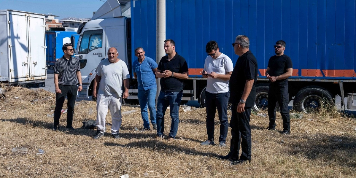 Açık havada, arkalarında büyük mavi ve beyaz tırların park halinde olduğu boş bir arazide duran, aralarında Başkan Eşki'nin de bulunduğu yedi kişilik erkek grubu. Gruptakiler günlük kıyafetler içinde, bir konu üzerinde konuşurken veya çevreyi incelerken görülüyor.