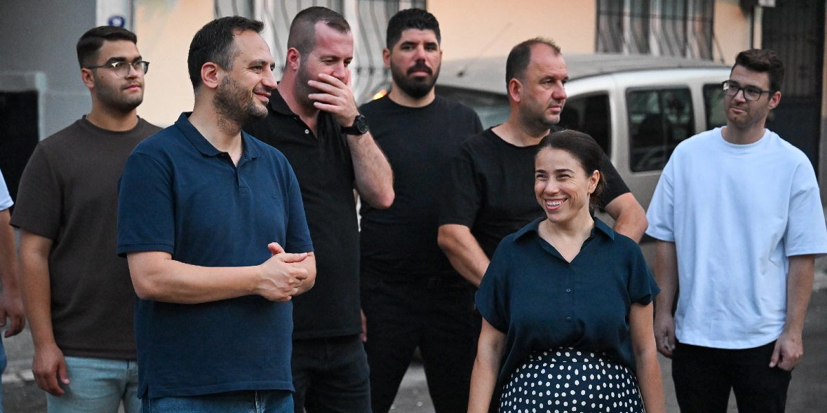 Açık havada, bir sokak ortamında bir araya gelmiş, aralarında Bornova Belediye Başkanı Ömer Eşki'nin de bulunduğu bir grup insan, bir noktaya bakarak sohbet ediyor. Ön planda lacivert polo yaka tişörtlü Ömer Eşki ve yanında lacivert bluz ile puantiyeli etek giymiş gülümseyen bir kadın yer alıyor. Arka planda ise siyah tişörtlü erkekler ve beyaz tişörtlü genç bir erkek bulunuyor.