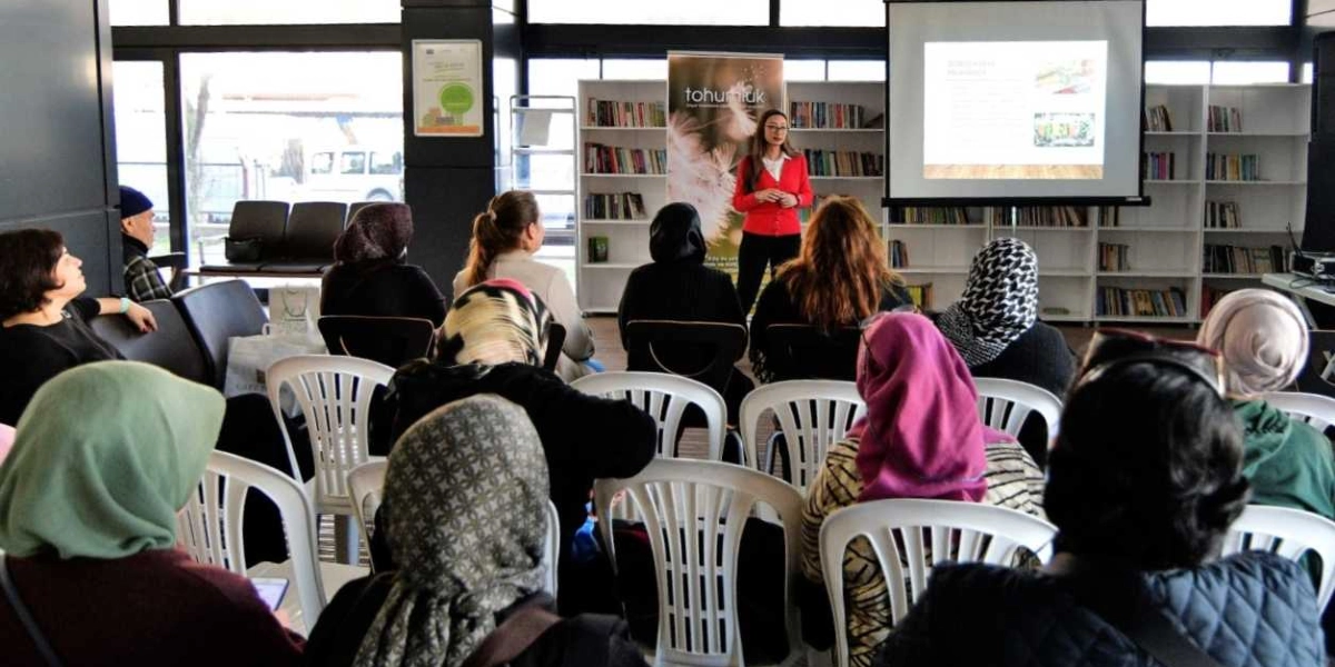 Kütüphane benzeri bir ortamda, beyaz plastik sandalyelerde oturan bir grup kadın, önlerinde sunum yapan bir konuşmacıyı dinliyor. Kırmızı hırkalı kadın konuşmacı, arkasındaki projeksiyon perdesinde yansıtılan bir sunum eşliğinde bilgi veriyor. Arka planda kitaplarla dolu raflar ve üzerinde 