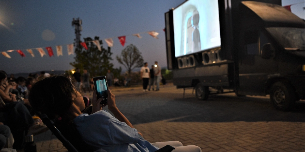 Alacakaranlıkta açık havada düzenlenen bir film gösteriminde, katlanır sandalyede oturan bir çocuk elindeki telefonla dev ekrana yansıtılan animasyon filmini kaydetmektedir. Arka planda, üzerine Türk bayrakları asılmış bir kamyonun yan tarafına monte edilmiş büyük bir ekran ve izleyiciler yer almaktadır.