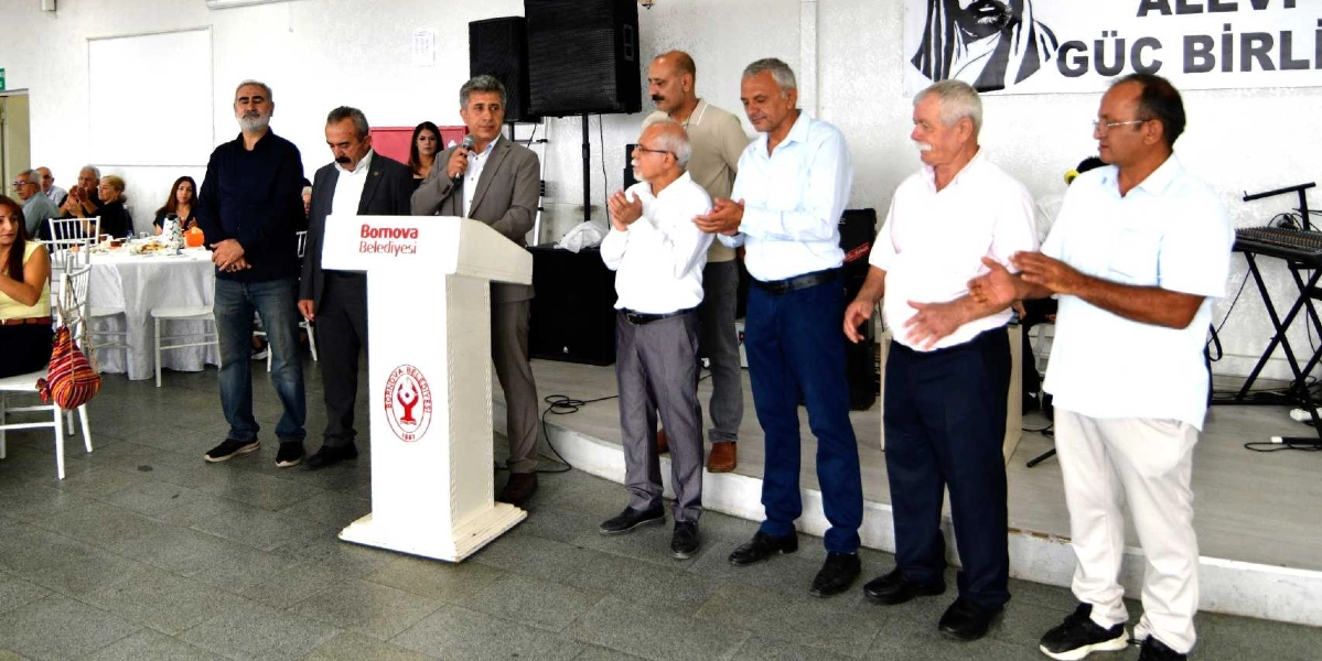 Bornova Belediyesi kürsüsünde konuşma yapan bir yetkili ve yanında duran Alevi Güçbirliği temsilcileri. Kürsüde Bornova Belediyesi logosu yer alırken, arka planda üzerinde 