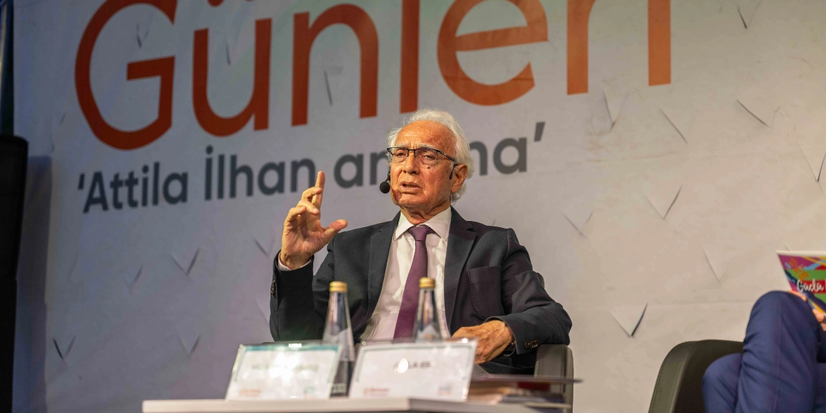 Beyaz saçlı, gözlüklü ve koyu renk takım elbiseli bir konuşmacı, arkasında 