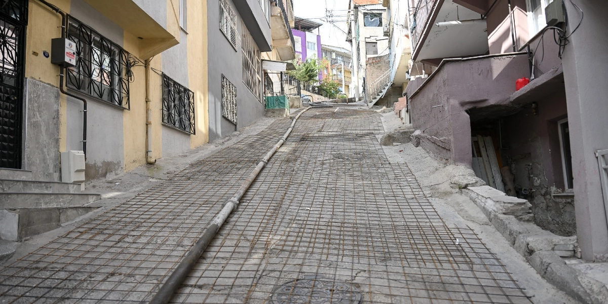 Bornova'da dik bir yokuşun zeminine beton dökülmeden önce yerleştirilmiş çelik hasır donatıları ve yolun ortasından geçen uzun bir boru hattı görülüyor. Yolun iki yanında çok katlı apartmanlar bulunuyor.
