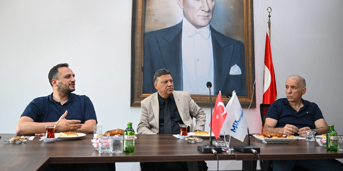 Üç erkek, üzerinde simit, çay ve maden suyu bulunan uzun bir toplantı masasında oturuyor. Ortada oturan kişi bej renkli bir ceket ve siyah gömlek giymiş, yanındakiler ise lacivert polo yaka tişört giymiş durumda. Masanın arkasındaki duvarda büyük bir Mustafa Kemal Atatürk portresi ve yanında Türk bayrağı yer alıyor. Masanın üzerinde ayrıca MTK yazılı küçük bir masa bayrağı bulunuyor.