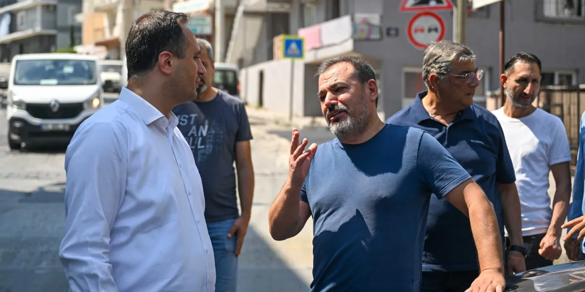 Belediye başkanı olduğu anlaşılan beyaz gömlekli bir kişi, sokakta yanındaki bir grupla sohbet ederken el hareketleriyle bir şeyler anlatıyor. Grubun geri kalanında farklı tişörtler giymiş erkekler bulunuyor ve arka planda bir trafik levhası ile beyaz bir ticari araç yer alıyor.