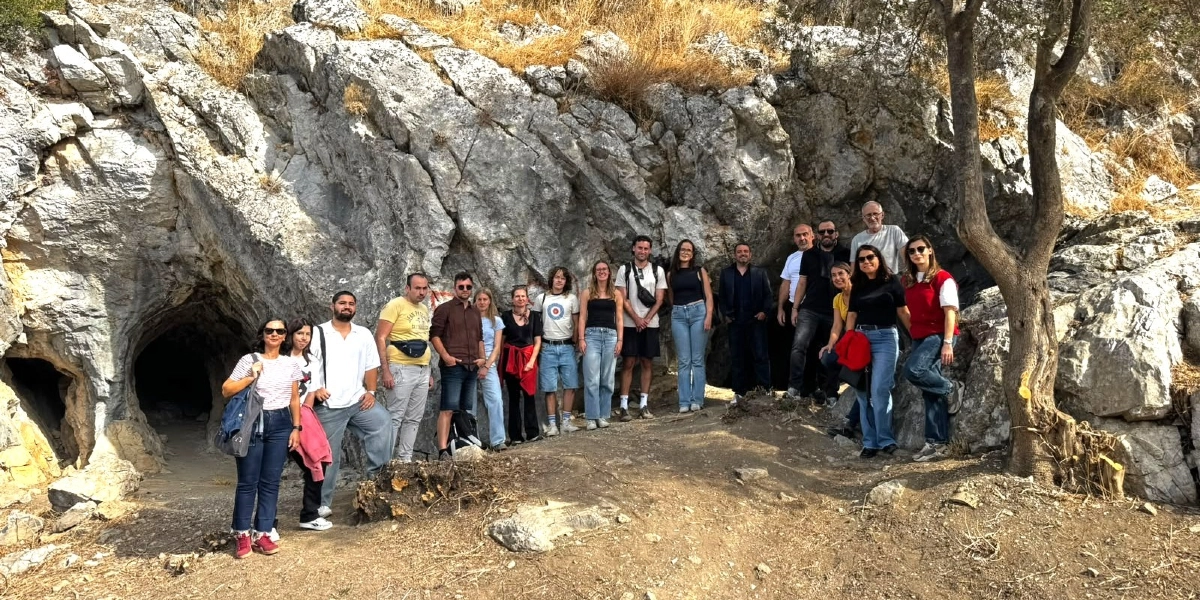 Bornova'daki tarihi bir mağaranın önünde yan yana dizilmiş, günlük kıyafetler içindeki yaklaşık yirmi kişilik bir grup insan, güneşli bir günde kameraya gülümseyerek poz veriyor. Arka planda gri renkli, kayalık bir yamaç ve sağ tarafta gövdesi belirgin bir ağaç yer alıyor.