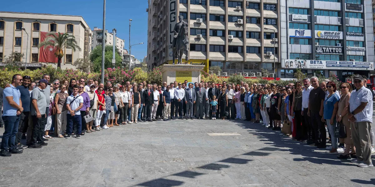 Bornova Cumhuriyet Meydanı'nda, Atatürk anıtının önünde toplanmış geniş bir kalabalık grup, 9 Eylül kutlamaları kapsamında yan yana dizilerek poz veriyor. Arka planda çok katlı binalar ve üzerinde Türk bayrağı asılı olan bir yapı görülüyor. Gün ışığında çekilen karede, farklı yaş ve giyim tarzlarından oluşan katılımcılar, meydanın merkezindeki atlı Atatürk heykeline dönük şekilde duruyor.