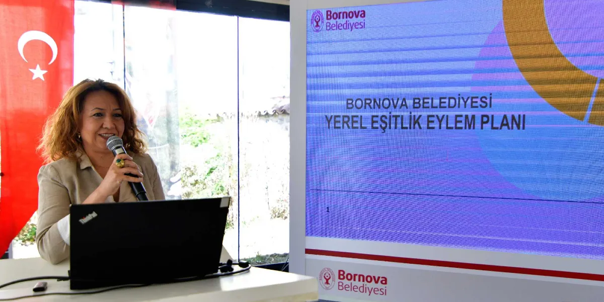 Kürsüde mikrofonla konuşma yapan, açık renkli ceket giymiş kumral bir kadın ve yanında üzerinde Bornova Belediyesi Yerel Eşitlik Eylem Planı yazılı büyük bir dijital ekran yer alıyor. Arka planda Türk bayrağı ve aydınlık bir pencere bulunuyor.