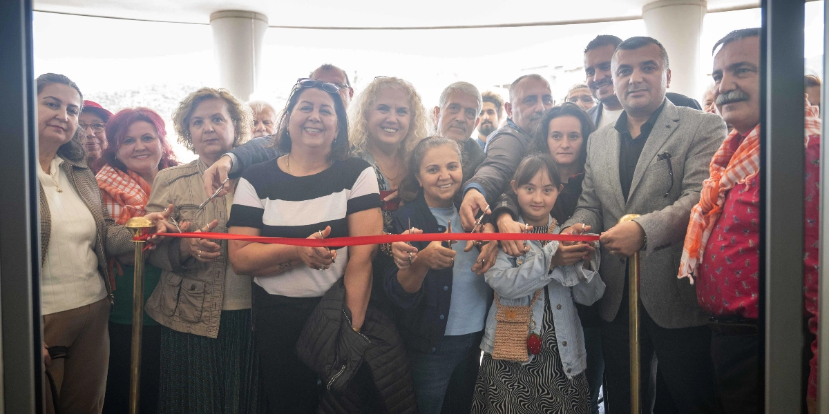 Bir grup kadın, erkek ve bir çocuk, ellerindeki makaslarla kırmızı bir açılış kurdelesini kesmek üzere bir araya gelmiş. Hepsi gülümseyerek kameraya bakıyor. Ortam aydınlık ve modern bir bina girişi gibi görünüyor.