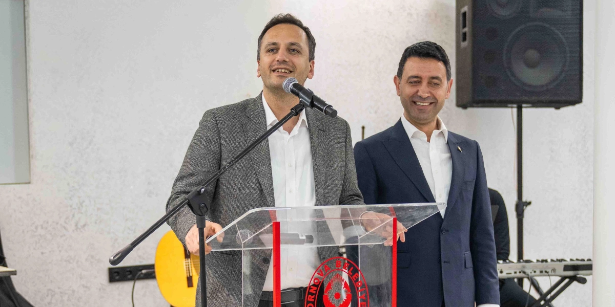 Kürsüde konuşma yapan gri ceketli bir erkek ve yanında gülümseyerek onu dinleyen lacivert takım elbiseli bir erkek yer alıyor. Kürsünün üzerinde Bornova Belediyesi logosu bulunurken, arka planda bir hoparlör ve kısmen görünen bir gitar ile klavye yer alıyor.