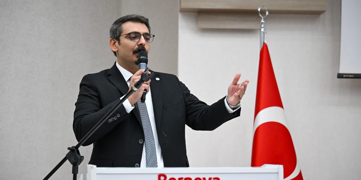 Siyah takım elbiseli, beyaz gömlekli ve gri kravatlı, gözlüklü bir erkek, üzerinde Bornova Belediyesi logosu bulunan bir kürsüde mikrofonla konuşma yapıyor. Konuşmacı bir elini mikrofonu tutmak için kullanırken diğer elini jest yaparak havaya kaldırmış durumda. Sağ tarafında ise bir Türk bayrağı yer alıyor.