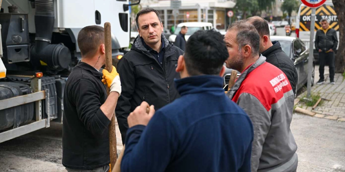 Bornova Belediye Başkanı, üzerinde Bornova Belediyesi Fen İşleri Müdürlüğü yazılı üniformalar giyen saha çalışanlarıyla bir şantiye alanında incelemelerde bulunuyor. Başkan siyah bir mont giymiş, işçilerle konuşurken dikkatle onları dinliyor. Arka planda bir iş makinesi ve trafik akışının olduğu bir cadde görülüyor.