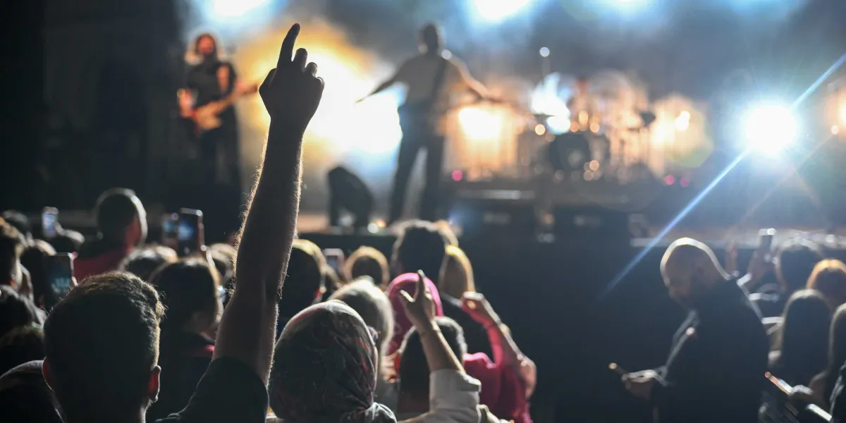 Sahne ışıkları altında konser veren bir müzik grubunu izleyen kalabalık bir izleyici kitlesi. Ön planda bir kişi elini havaya kaldırmış, diğer izleyiciler ise ellerinde telefonlarla sahneyi kaydediyor. Sahnedeki müzisyenler parlak ışıkların yarattığı silüetler halinde görünüyor.