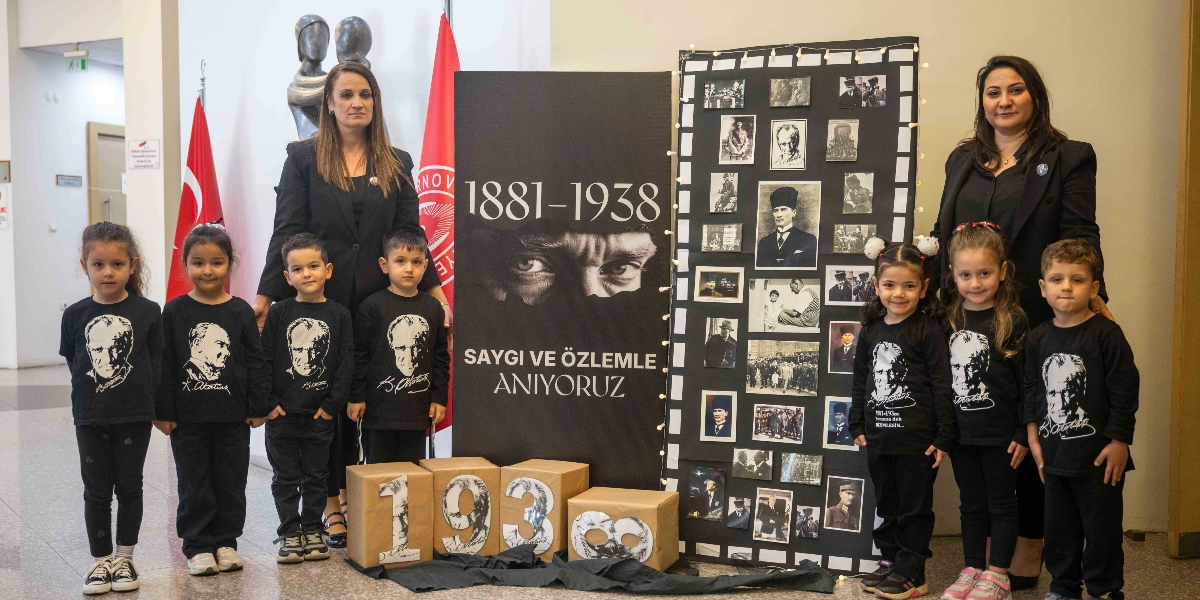 Atatürk'ü anma etkinliğinde, üzerinde Atatürk baskılı siyah tişörtler giyen yedi anaokulu öğrencisi, iki öğretmenleri ile birlikte poz veriyor. Grubun arkasında, üzerinde 