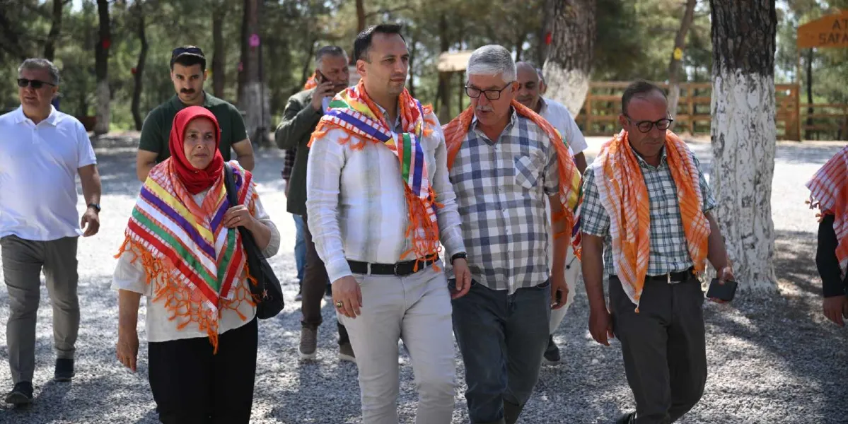 Açık havada, ağaçlık bir alanda yürüyen bir grup insan. Önde, omuzlarında renkli, yöresel desenli şallar bulunan bir kadın ve iki erkek yan yana yürüyor. Kadın kırmızı bir başörtüsü takıyor, yanındaki erkeklerden biri beyaz gömlek, diğeri ise kareli gömlek giyiyor. Grubun arkasında ve çevresinde yürüyüşe eşlik eden başka erkekler de bulunuyor. Arka planda ağaçlar ve ahşap bir çit yapısı görülüyor.