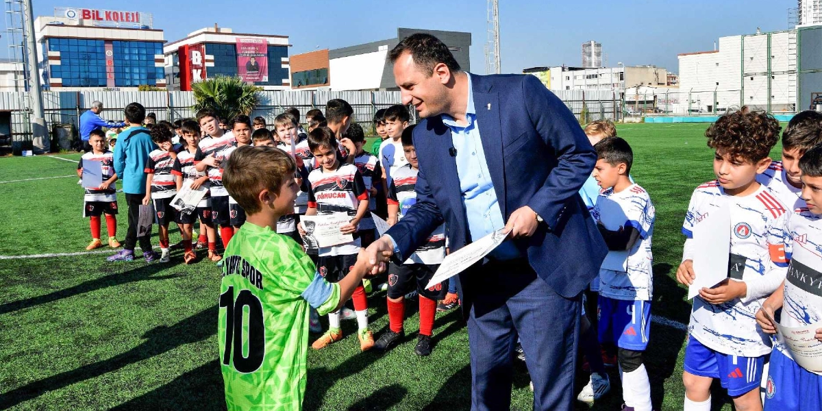 Açık havada, suni çim kaplı bir futbol sahasında, lacivert takım elbiseli bir yetişkin, üzerinde 10 numara yazılı yeşil bir forma giyen küçük bir futbolcuyla el sıkışarak ona bir başarı belgesi veriyor. Çevrede, ellerinde benzer belgeler tutan, farklı renklerde formalar giymiş çok sayıda çocuk futbolcu, tören havasında bir sırada bekliyor. Arka planda ise Bil Koleji yazılı bir bina ve şehir yerleşimi görülüyor.