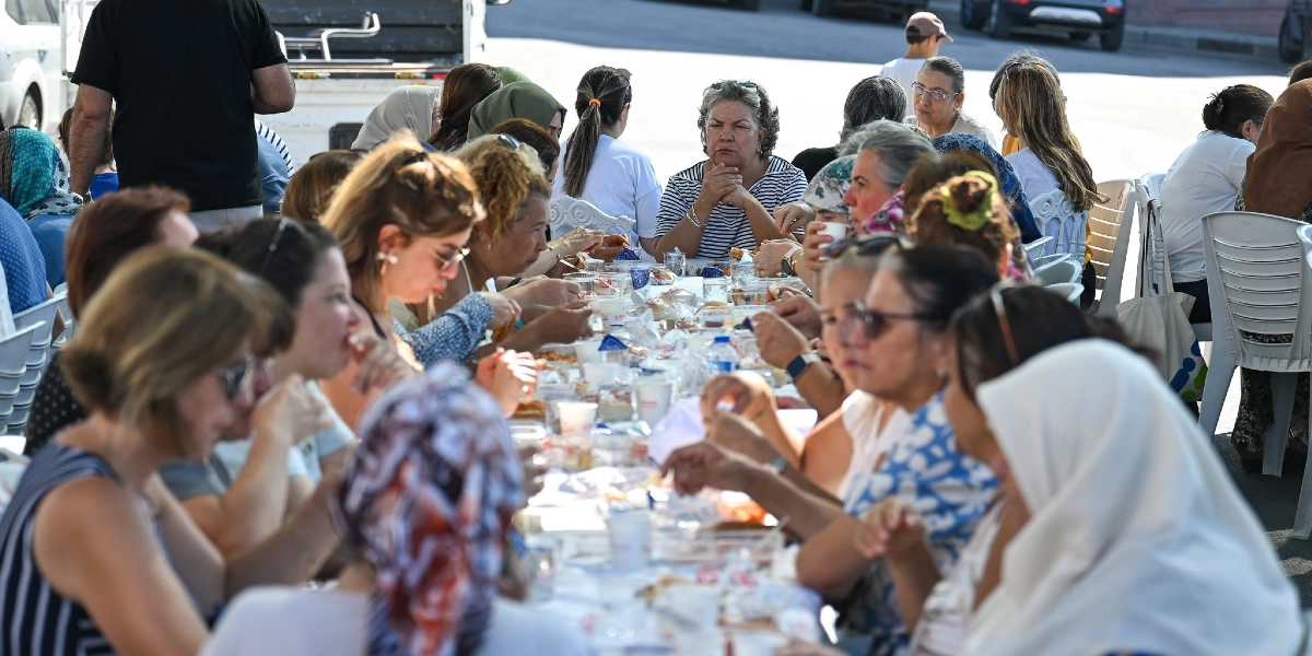 Açık havada uzun bir masada toplanmış, yemek yiyen ve sohbet eden kadınlar grubu. Masanın üzerinde çeşitli yiyecekler ve içecekler bulunuyor. Arka planda ise ayakta duran bir erkek ve park halindeki araçlar görülüyor.
