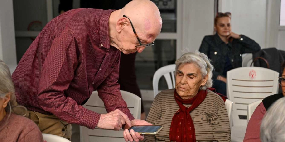 Bordo renkli gömlek giyen yaşlı bir adam, elindeki akıllı telefonu oturmakta olan yaşlı bir kadına göstererek bir şeyler anlatıyor. Kadın dikkatle telefona bakarken, arka planda başka katılımcılar da onları izliyor.