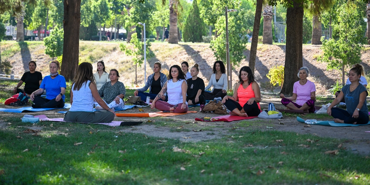 Ağaçlık bir park alanında, yoga matları üzerinde bağdaş kurmuş şekilde oturan ve meditasyon yapan bir grup kadın. Kadınlar farklı renklerde spor kıyafetleri giymiş, gözleri kapalı ve elleri dizlerinin üzerinde dinlenme pozisyonunda duruyorlar. Arka planda yeşil ağaçlar ve hafif eğimli bir toprak zemin görülüyor.