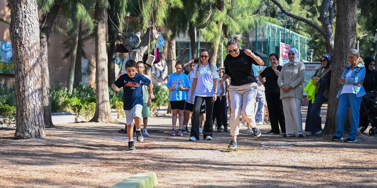 Ağaçlık bir park alanında, yere dizilmiş renkli halkaların üzerinde spor yapan bir çocuk ve ona eşlik eden bir kadın eğitmen görülüyor. Çevredeki diğer çocuklar ve yetişkinler, etkinliği izleyerek onlara destek veriyor.