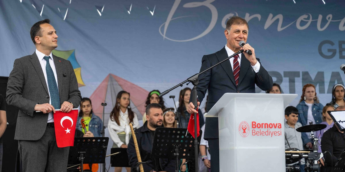 Bornova Belediye Başkanı, üzerinde Bornova Belediyesi logosu bulunan bir kürsüde mikrofonla konuşma yapıyor. Yanında elinde Türk bayrağı tutan bir yetkili bulunuyor. Arka planda ise müzik aletleriyle sahne alan bir grup çocuk ve genç yer alıyor.