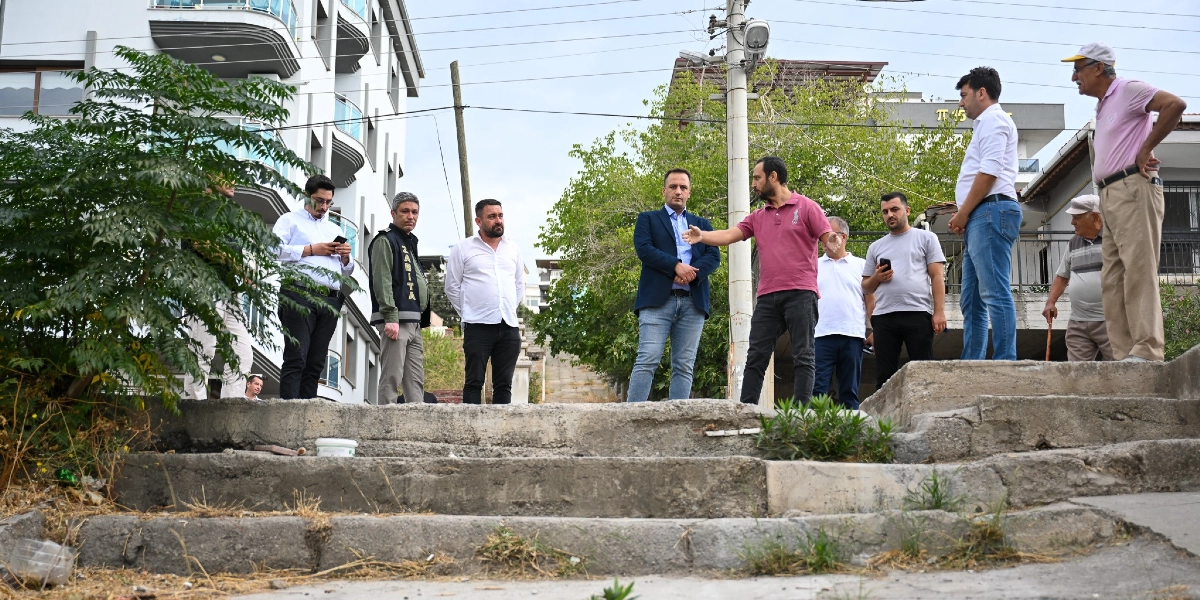 Bornova'da bir grup yetkili, yenilenmesi planlanan eski ve yıpranmış beton merdivenlerin başında incelemelerde bulunuyor. Kırmızı tişörtlü bir kişi eliyle merdiven alanını işaret ederek açıklama yaparken, çevresindeki diğer yetkililer ve zabıta personeli onu dinliyor. Arka planda modern apartmanlar ve ağaçlar yer alıyor.