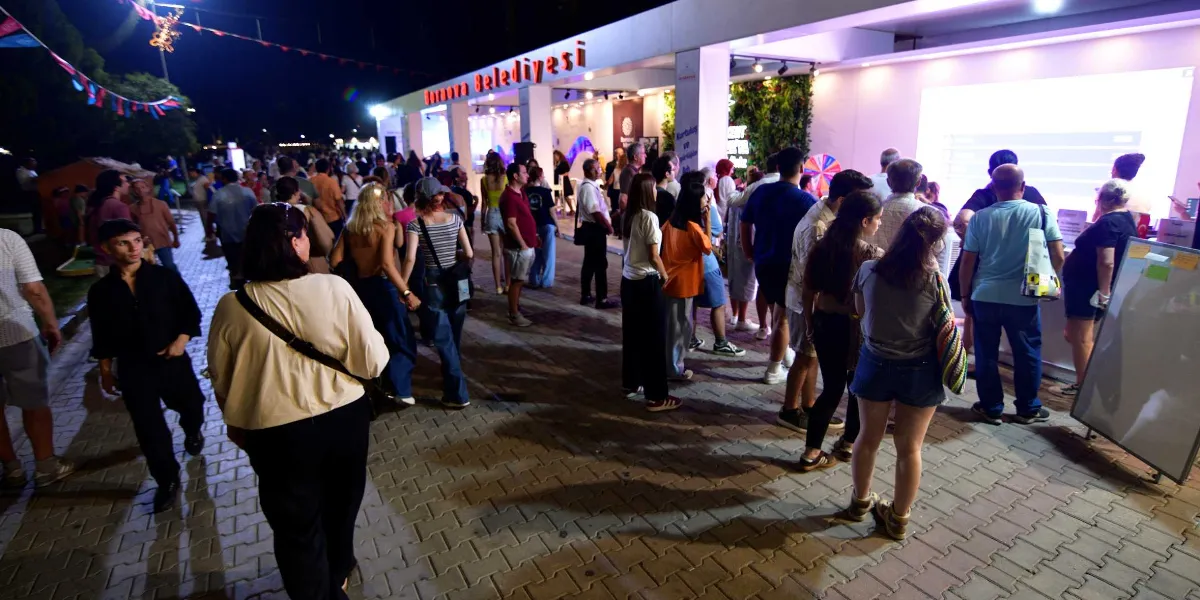 Gece vakti, üzerinde kırmızı harflerle Bornova Belediyesi yazan modern bir standın önünde toplanmış kalabalık bir grup insan. İnsanlar standın önündeki büyük ekranı izliyor, bazıları ise stant çevresinde sohbet ediyor. Alan, ışıklandırmalar ve asılı süslemelerle aydınlatılmış, parke taşlı bir yürüme yolunda hareketlilik yaşanıyor.