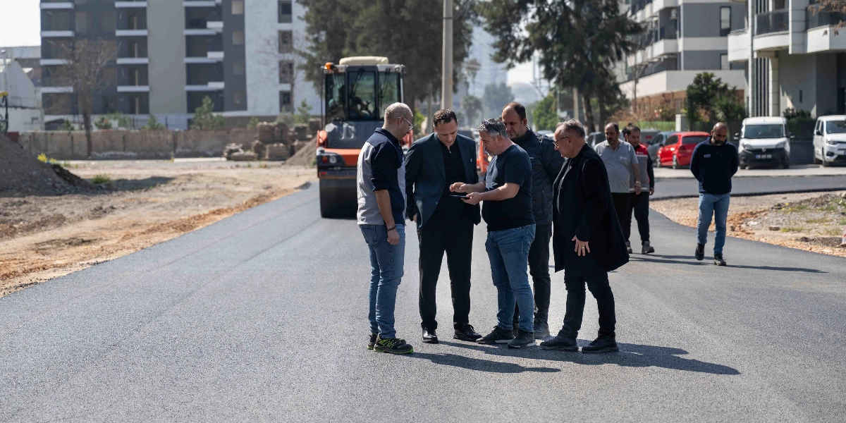 Yeni asfaltlanmış bir yol üzerinde duran bir grup yetkili, ellerindeki tablete bakarak çalışmaları inceliyor. Arka planda bir silindir iş makinesi ve çevredeki binalar görülüyor.