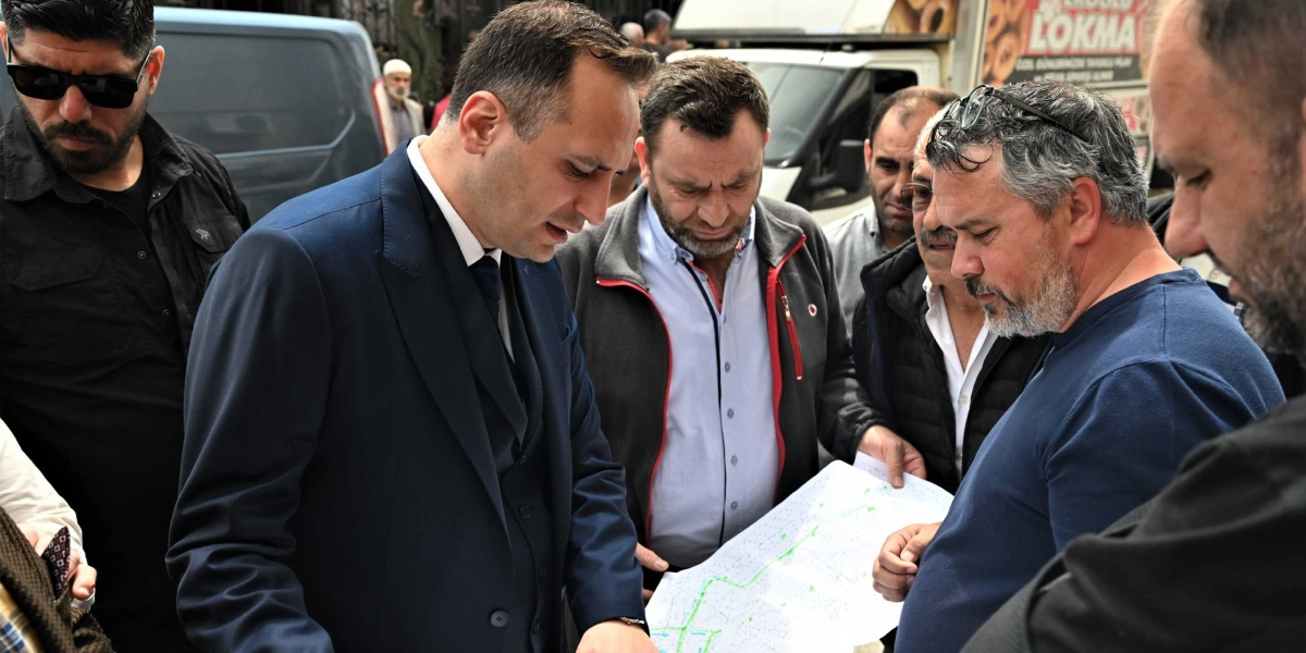 Lacivert takım elbiseli bir erkek, çevresindeki bir grup insanla birlikte ellerinde tuttukları bir haritayı veya proje planını inceliyor. Dış mekanda çekilen bu karede, grup üyeleri dikkatle kağıda bakarken, arka planda bir lokma aracı ve diğer insanlar yer alıyor.